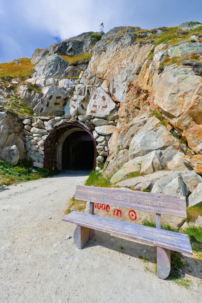our-services-1 A scenic view of an entrance to a mountain tunnel in Fiesch, Switzerland, showing rocky terrain and a wooden bench.